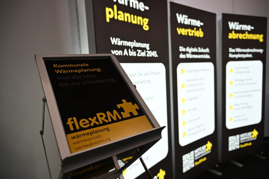 Ein schwarzer Info-Aufsteller von flexRM mit der Aufschrift "Kommunale Wärmeplanung: Von der Idee bis zur Umsetzung" vor aufgestellten Messe-Rollups.